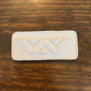 Puede incluir: Un parche de cuero blanco con un borde cosido. El parche presenta un diseño "W" estilizado en relieve en el centro. El parche es rectangular y tiene una superficie texturizada.