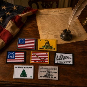 May include: A collection of embroidered patches with patriotic and historical themes. Designs include the American flag, the Gadsden flag, and phrases like "Join, or Die" and "An Appeal to Heaven." The patches are displayed on a wooden surface.
