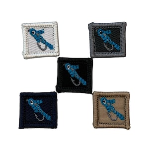 Può includere: Cinque toppe quadrate ricamate con un disegno di mosca da pesca blu. Le toppe sono in vari colori: bianco, nero, blu navy, grigio e beige. Ogni toppa presenta una mosca blu dettagliata con un amo curvo.