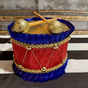 May include: A decorative drum ornament featuring a red fabric body with blue ruffled trim at the top and bottom. The drum is accented with gold braided trim, small gold studs, and thin gold cord lacing. Two gold glitter drumsticks rest on top.