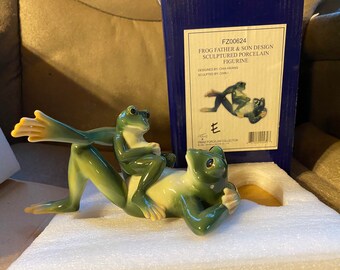May include: A porcelain figurine of a frog father and son. The father frog is lying on its back with its legs outstretched. The son frog is sitting on the father's back with its arms around the father's neck. The figurine is green and white and has a detailed design. The box reads "FZ00624 Frog Father & Son Design Sculptured Porcelain Figurine Designed by Chia Heiang Sculptured by Chini Franz Porcelain Collection"