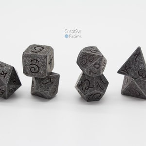 Könnte beinhalten: Set mit sechs grauen, steintexturierten Polyederwürfeln für Tabletop-Spiele. Jeder Würfel hat schwarze Zahlen. Das Set enthält einen d4, d6, d8, d10, d12 und d20. Das Creative Realms Logo ist im Hintergrund.