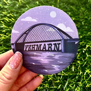 Puede incluir: Una pegatina circular de color morado y blanco con un gráfico de un puente y la palabra "FEHMARN" en texto negro. La pegatina está sobre una superficie verde de hierba.