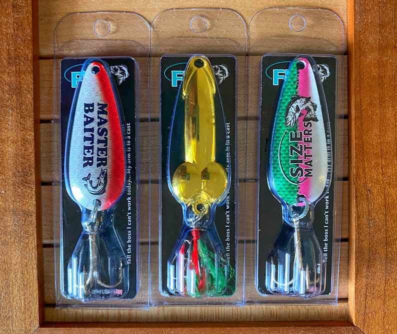 May include: Three fishing lures with humorous sayings on them. The lures are packaged in clear plastic and have a silver, gold, and green and pink color scheme. The sayings on the lures are "Master Baiter", "Size Matters", and "Tell the boss I can't work today... My arm is in a cast".