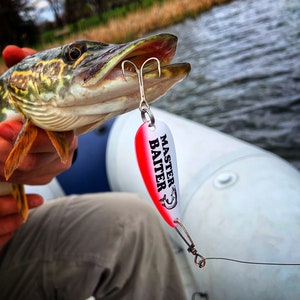 May include: A close-up of a pike fish being held by a person. The fish is being held by the mouth and is facing the camera. A red and white fishing lure with the text "Master Baiter" is attached to the fish's mouth.