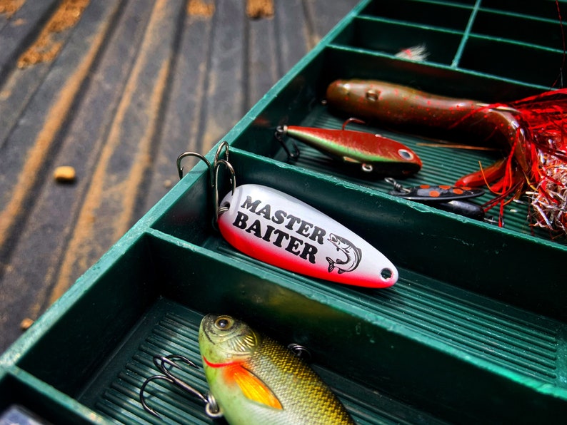 May include: A green tackle box with various fishing lures, including a white and red lure with the text "MASTER BAITER" printed on it.