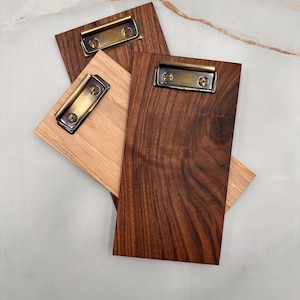 May include: Three wooden clipboards with brass clasps. The clipboards are stacked on top of each other. The top clipboard is dark brown wood, the middle clipboard is light brown wood, and the bottom clipboard is dark brown wood.