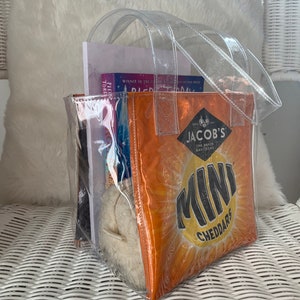 May include: A clear plastic tote bag with two handles. The bag contains a book, a white stuffed animal, and a bag of Jacob's Mini Cheddars.