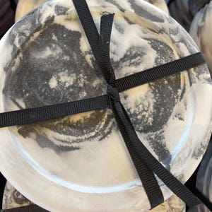 May include: A circular, flat object with a marbled pattern in shades of white, gray, and black. The surface has a smooth, polished appearance. A thin, black fabric ribbon is tied in a simple knot across the center of the circular object.