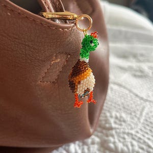 Peut inclure: Un charm en forme de canard perlé, avec une tête verte, un corps marron et blanc, et des pieds orange. Le charm est attaché à un fermoir doré, suspendu à un sac en cuir marron. Le canard a un petit accent rouge sur la tête.