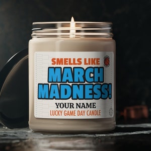 May include: A brown candle with a white label that says "Smells Like March Madness!" and "Your Name Lucky Game Day Candle". The label also features a basketball graphic.