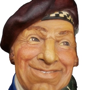 May include: A ceramic bust of a man wearing a burgundy beret with a black and white checkered band, a blue shirt, and a green, red, and yellow plaid scarf.