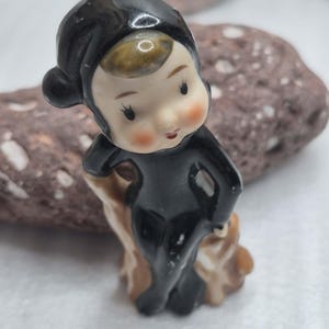 May include: A small ceramic figurine of a character wearing a black hooded suit and a black cap. The figure has a pale face with rosy cheeks, brown hair peeking from under the hood, and is posed sitting on a brown, textured tree stump base.