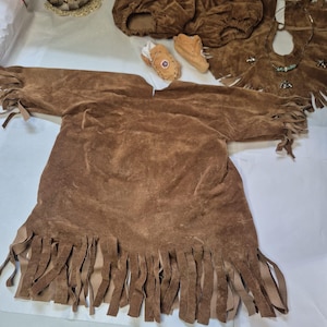 Op de afbeelding: Een bruine suède kostuumset, inclusief een tuniek met franjes, een broek, een vest en mocassins. De set heeft een Indiaans thema. Er is ook een uilbeeldje aanwezig.
