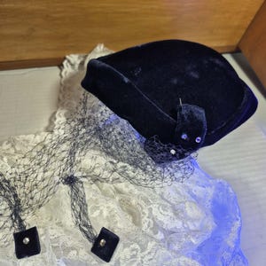 May include: A navy blue velvet hat with a matching veil and decorative accents. The hat is square-shaped and features a small, embellished flap. The veil is black and attached to the hat with small, square velvet pieces.
