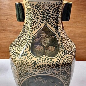 May include: A decorative ceramic vase with a cream-colored base and dark green accents. The vase features intricate floral and vine patterns, with a heart-shaped design in the center. Two dark green handles are on the sides. The vase is approximately 12 inches tall.