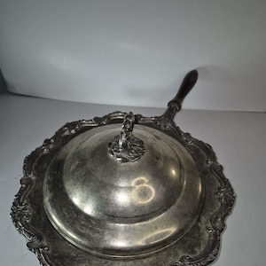 Puede incluir: Mantequera antigua plateada con tapa y mango de madera oscura. El plato tiene un borde festoneado adornado y un asa decorativa en la tapa. El acabado plateado muestra signos de edad y uso.
