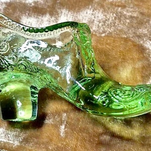 Puede incluir: Una zapatilla decorativa de cristal verde con intrincados detalles florales y de volutas. El zapato de estilo vintage presenta un tacón pequeño y una punta puntiaguda, perfecto para exhibir o como objeto de colección único.