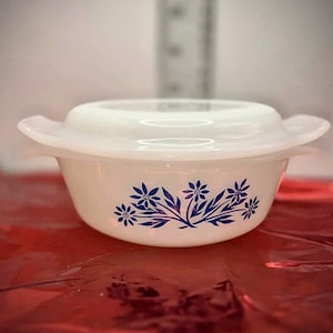 Puede incluir: Un cuenco blanco con tapa, con un diseño floral azul. El cuenco tiene dos asas y probablemente está hecho de vidrio o cerámica. Se muestra sobre una superficie roja. El diseño del cuenco sugiere un estilo vintage.