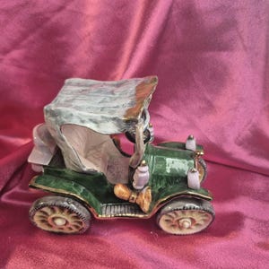 May include: A ceramic antique car figurine in shades of green, gray, and beige. The vintage car has a convertible top and detailed wheels. The background is a deep pink fabric.