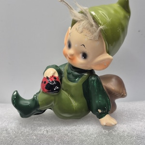 Puede incluir: Una figurita de elfo de cerámica verde con pelo blanco, sentada en un hongo marrón. El elfo sostiene una mariquita roja y negra en su mano.
