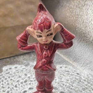 Könnte beinhalten: Eine kleine, glänzende, dunkelrote Keramikfigur eines elfenartigen Wesens mit spitzem Hut, einer Jacke mit Kragen und drei Knöpfen sowie spitzen Schuhen. Die Figur hat große Ohren, einen lächelnden Gesichtsausdruck und erhobene Hände.