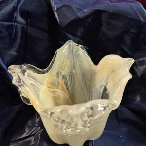 May include: A clear glass bowl with a unique, wavy shape. The bowl has a creamy white and yellow marbled design throughout. The edges of the bowl are clear and have a slightly ruffled appearance. The bowl is sitting on a dark blue surface.