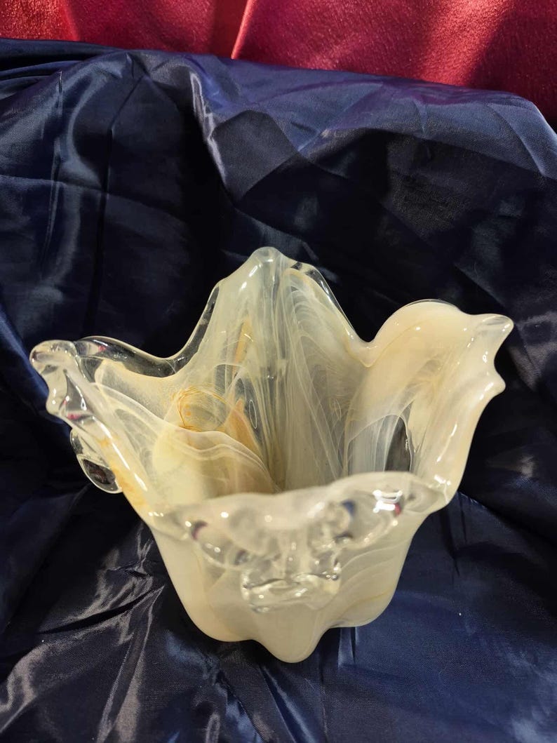 May include: A clear glass bowl with a unique, wavy shape. The bowl has a creamy white and yellow marbled design throughout. The edges of the bowl are clear and have a slightly ruffled appearance. The bowl is sitting on a dark blue surface.