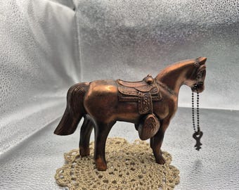 May include: A copper-toned horse figurine featuring a detailed saddle and bridle. A small metal ball chain with a decorative key charm hangs from the horse's head. The horse stands in a profile position, showcasing its textured mane and tail.