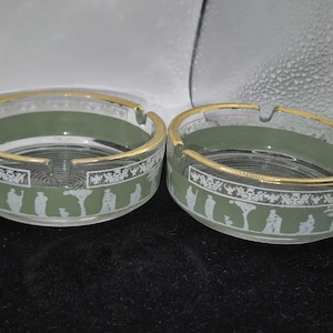 May include: Two clear glass ashtrays with a gold rim and a green band. The ashtrays have a white decorative pattern of figures and trees. The ashtrays are round and have a small notch for cigarettes.