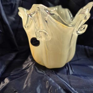 May include: A handcrafted, off-white and clear glass vase with a unique, ruffled design. The vase has a flared opening and a textured surface. A small, black label with white text is affixed to the side. The vase is likely for decorative purposes.