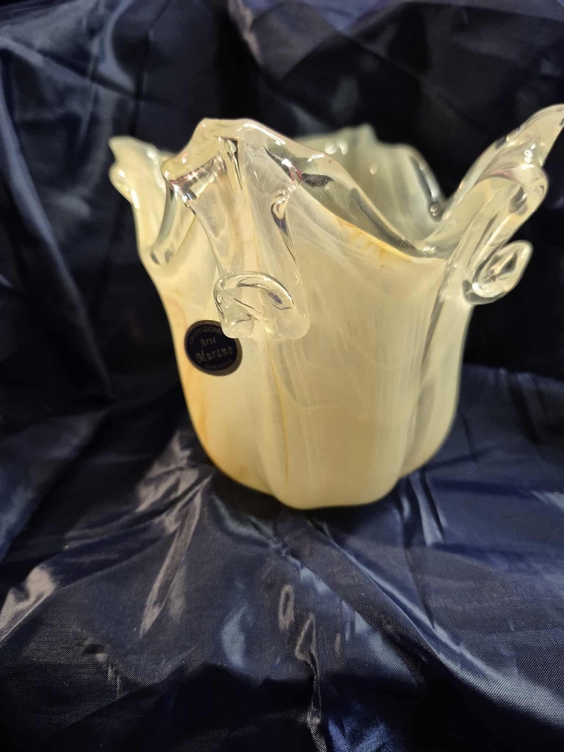 May include: A handcrafted, off-white and clear glass vase with a unique, ruffled design. The vase has a flared opening and a textured surface. A small, black label with white text is affixed to the side. The vase is likely for decorative purposes.