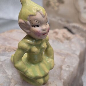 May include: A small ceramic figurine of a kneeling elf wearing a yellow pointed hat and a matching yellow dress. The elf has a pale face with painted black eyes, arched eyebrows, and rosy pink cheeks, with pointed ears extending from the sides.