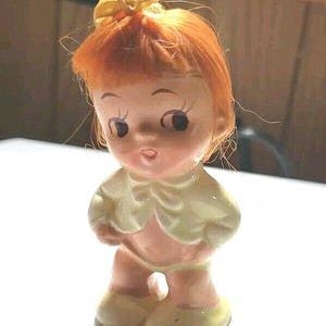 Può includere: Una piccola statuetta in ceramica di una bambina con i capelli rossi, che indossa un fiocco giallo, una giacca bianca e pantaloni rosa. La statuetta ha un'espressione dolce e innocente.