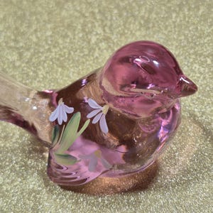 May include: A pink glass bird figurine with painted white flowers and green leaves. The bird has a clear tail and is sitting on a gold surface. The bird is a decorative item.