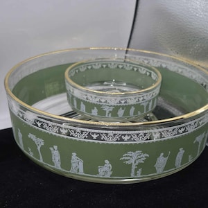 May include: A two-piece glass serving set with a green interior and gold trim. The exterior features a white decorative pattern with figures and trees. The set includes a large bowl and a smaller bowl that fits inside.