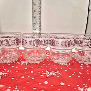 May include: Four clear glass tumblers with a decorative band featuring a profile portrait in a reddish-brown color. The glasses are on a red surface with white snowflake patterns.