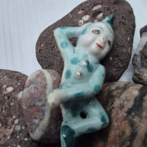 May include: A small porcelain figurine of a jester wearing a light blue suit with dark blue polka dots. The jester has a pointed hat, a painted face with red lips, and is posed with one arm raised behind the head and one leg bent at the knee.
