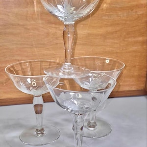 Op de afbeelding: Een set van vier heldere champagne coupe glazen. Elk glas heeft een brede kom met geëtste bloemmotieven en een slanke steel. De glazen zijn in een gestapelde formatie gerangschikt, wat hun elegante ontwerp benadrukt.