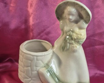 Puede incluir: Una jardinera de cerámica vintage con forma de mujer con vestido y sombrero, sosteniendo flores. La jardinera tiene un recipiente separado en forma de pozo. La combinación de colores es principalmente blanquecina con detalles verdes.