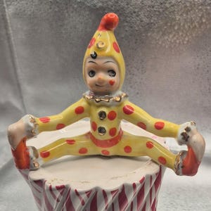May include: A ceramic figurine of a jester in a yellow suit with red polka dots, wearing a matching pointed hat. The jester is in a seated position with legs extended outwards, featuring a ruffled collar and red accents on the feet and face.