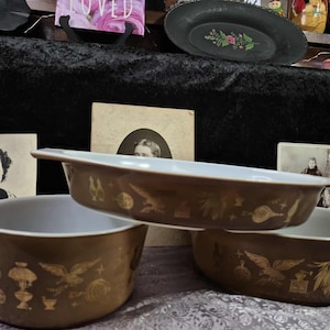 Może przedstawiać: Trzy brązowe naczynia do zapiekania Pyrex z białym wnętrzem i złotymi wzorami dekoracyjnymi. Naczynia są w różnych rozmiarach i ułożone w grupie. Tło obejmuje zdjęcia vintage i przedmioty dekoracyjne.