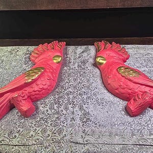 May include: Two matching, vibrant pink parrot wall decorations with gold accents. The parrots are perched on wooden branches, with detailed feathering and crests. These decorative pieces add a tropical flair to any room.