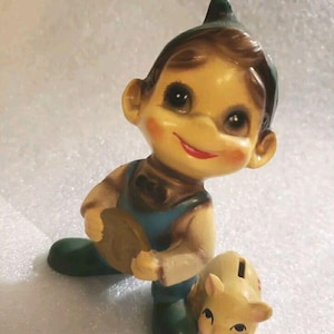 Pode incluir: Estatueta de cerâmica vintage de um menino ajoelhado segurando uma moeda, com um pequeno cofrinho em forma de gato. O menino tem bochechas rosadas, um chapéu verde e macacão azul. O banco de gatos é amarelo.