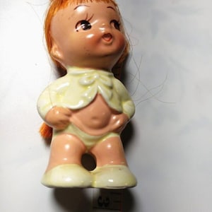 May include: A small ceramic doll with a yellow dress and red hair. The doll has a sweet expression and is wearing white shoes.