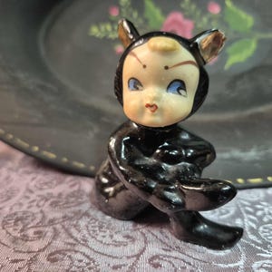Peut inclure: Figurine vintage d'une figurine noire assise en forme de chat avec un visage humain. La figurine a les yeux bleus, une petite bouche rouge et porte une tenue noire avec des oreilles de chat. La figurine est sur une surface à motifs avec une assiette sombre en arrière-plan.