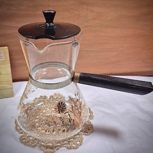 Könnte beinhalten: Eine Vintage-Kaffeekanne aus Glas mit schwarzem Deckel und Griff. Der transparente Glaskörper weist ein goldenes Blumendesign auf und steht auf einer goldenen gehäkelten Untersetzer. Die Kanne ist für den Gebrauch auf dem Herd konzipiert.