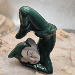 May include: A glossy dark green ceramic figurine of a pointed-ear elf in a contorted, arched pose. The elf has a pale face with painted black eyes, arched eyebrows, and a small red mouth, with its body bent backward in a flexible, acrobatic shape.
