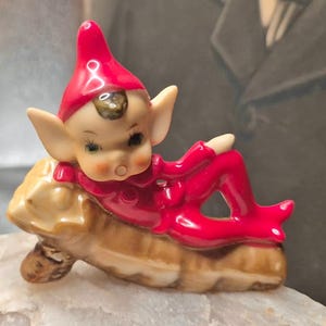 May include: A ceramic figurine of a small elf with a pointed red hat and red clothing, reclining on a textured brown log. The elf has large ears, painted facial features, and a glossy finish. The figure is depicted in a resting pose on the log.