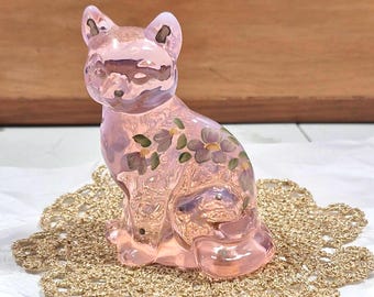 May include: A pink glass cat figurine with floral accents in purple and green. The cat is sitting on a gold crocheted doily. The figurine has a glossy finish and is set against a wooden background.